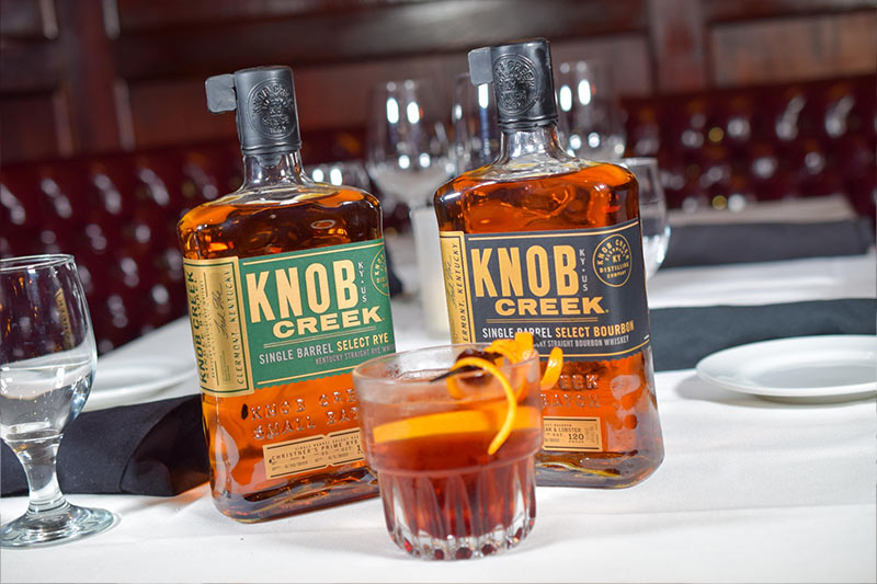 two bottles of Knob Creek whiskey next to a poured glass of whiskey with orange peels in it.