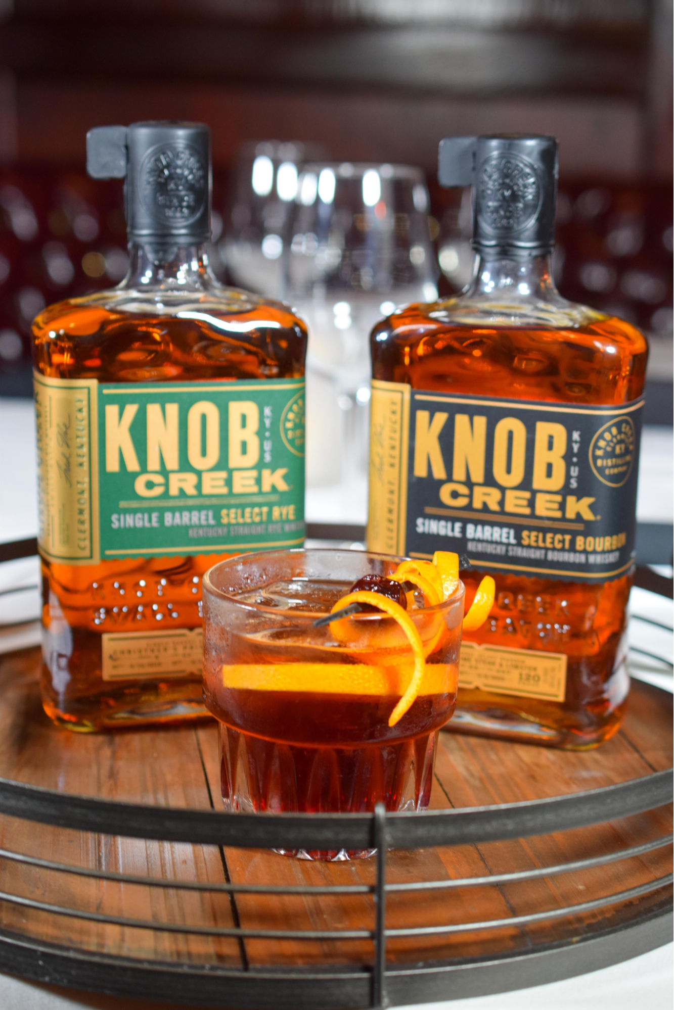 Knob Creek whiskey prepared in a glass with a cherry and orange peel. Two bottles of Knob Creek whiskey behind the glass.