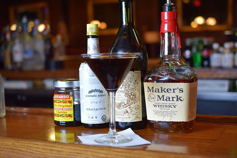 Chrisnter’s Manhattan in a martini glass with the different bottles of liquor and ingredients in the background.